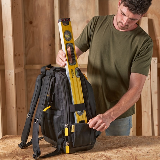 Photo of STANLEY FMST1-80144 black tool backpack on a workbench, with a yellow level being placed inside front pocket.