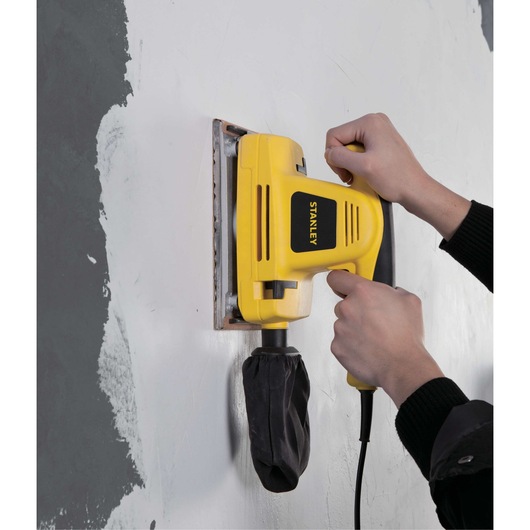 Stanley SSS310 yellow electric sander with dust bag sanding a wall.