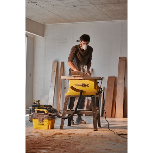 STANLEY SST2001-B2 vacuum cleaner connected to table saw in workshop with wooden planks in background, side angle.
