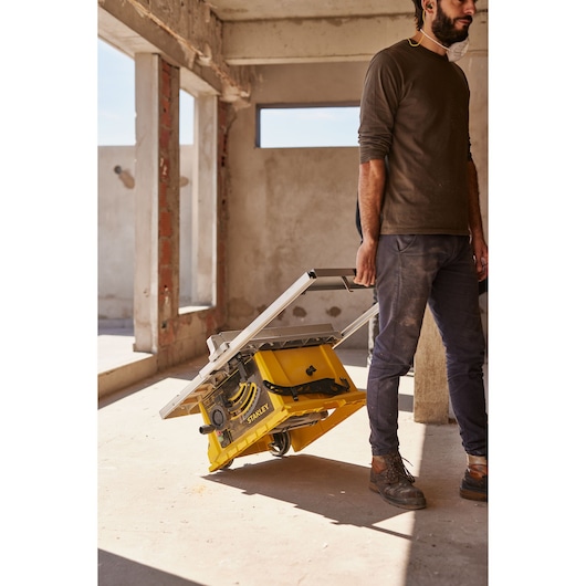 Photo of a person transporting a STANLEY SST2001-B2 table saw with wheels at a construction site.