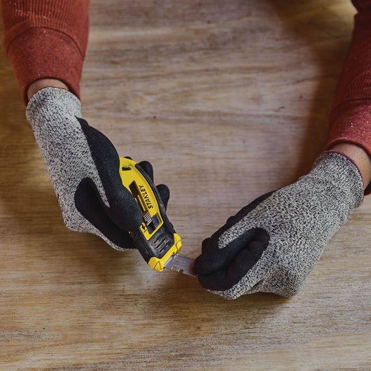 Photo of STANLEY STHT10432 utility knife with yellow and black handle, being held by gloved hands on a wooden surface.