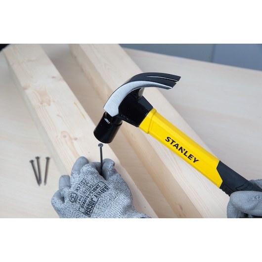 STANLEY STHT51391 claw hammer with yellow and black handle used to drive a nail into wood by a gloved hand.