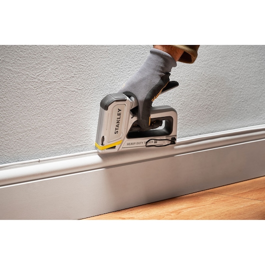 STANLEY STHT70250 heavy duty stapler in use, gloved hand pressing tool against white baseboard, angled side view.