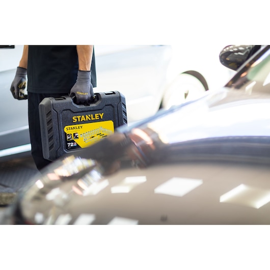 Photo of a person holding a STANLEY STMT82832-LA black hard tool case with yellow label, automotive setting.