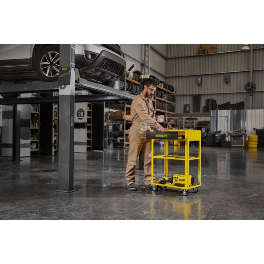 STANLEY STST98520LA yellow tool cart with three shelves in a garage, mechanic standing beside cart.